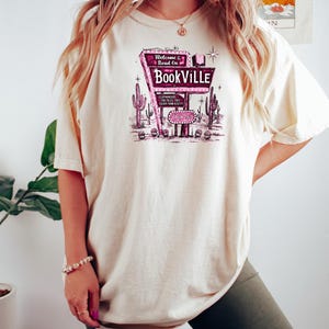 May include: A woman wearing a white t-shirt with a pink and white graphic of a retro motel sign that says "Welcome & Read On Bookville". The sign has a star and a book on it. The woman is wearing green biker shorts.