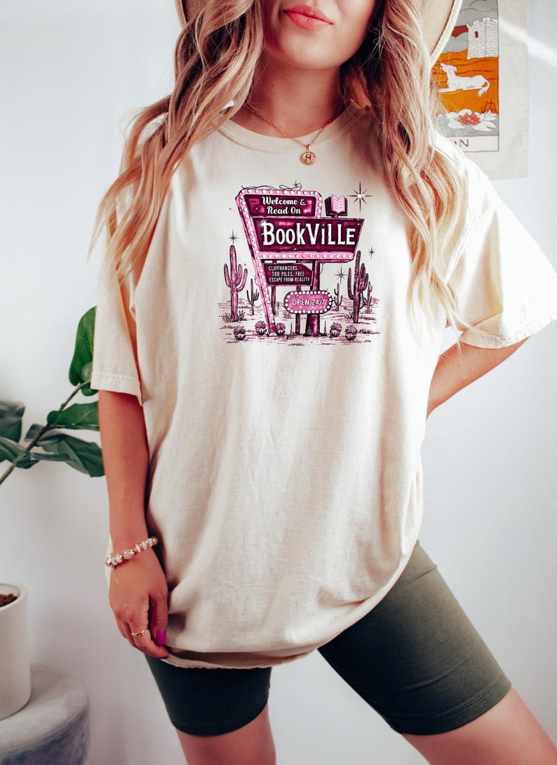 May include: A woman wearing a white t-shirt with a pink and white graphic of a retro motel sign that says "Welcome & Read On Bookville". The sign has a star and a book on it. The woman is wearing green biker shorts.