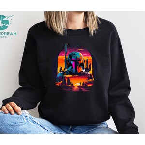 Pu&ograve; includere: Una felpa nera con una grafica colorata di un personaggio di Star Wars, Boba Fett, in piedi in un paesaggio desertico al tramonto. La grafica &egrave; in stile retr&ograve; con colori vivaci e dettagli pixelati.