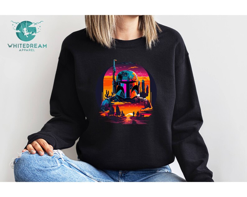 Pu&ograve; includere: Una felpa nera con una grafica colorata di un personaggio di Star Wars, Boba Fett, in piedi in un paesaggio desertico al tramonto. La grafica &egrave; in stile retr&ograve; con colori vivaci e dettagli pixelati.