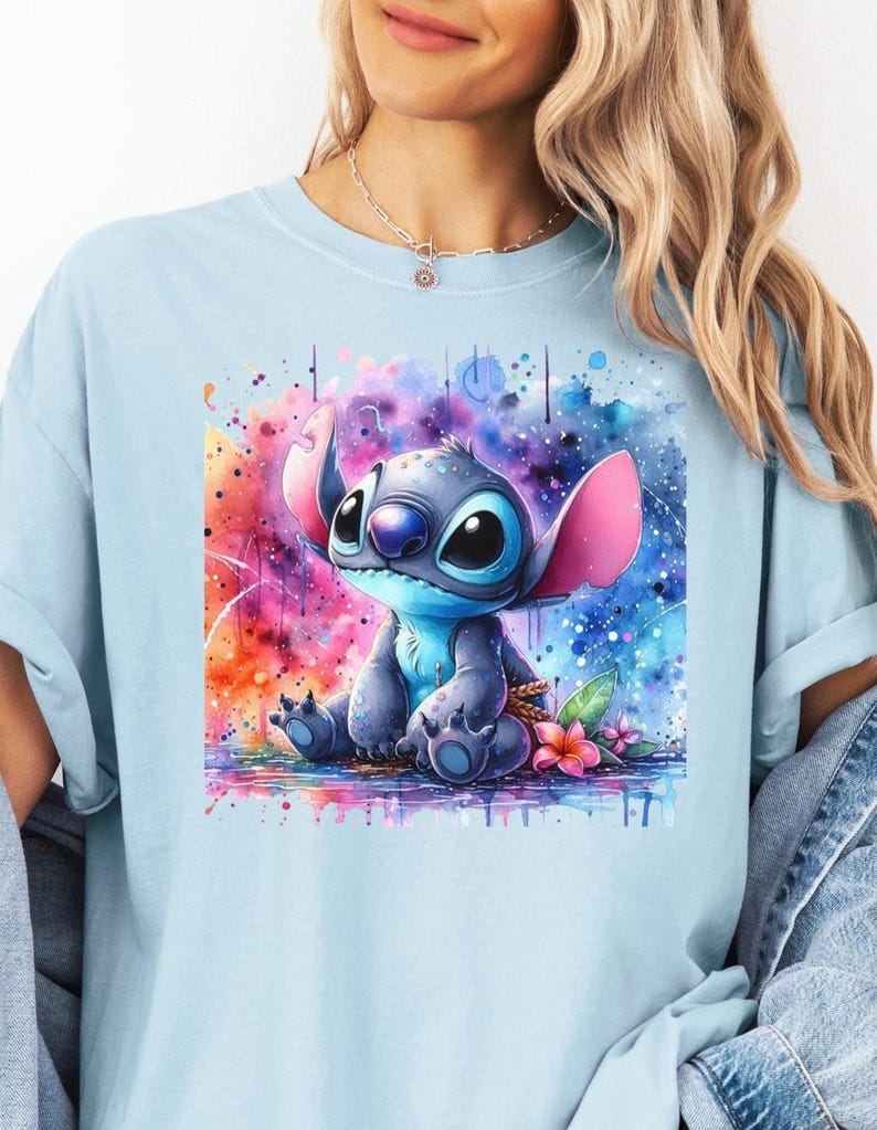 Pu&ograve; includere: T-shirt azzurra con grafica ad acquerello di Stitch di Lilo & Stitch. L'illustrazione mostra Stitch seduto su uno sfondo colorato di rosa, viola, blu e arancione. Fiori rosa sono in basso.