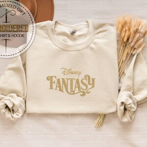 Peut inclure: Sweat-shirt cr&egrave;me avec l'inscription "Disney Fantasy" en lettres dor&eacute;es. Le sweat-shirt a des manches longues et un col rond. L'image comprend &eacute;galement un bouquet de bl&eacute; s&eacute;ch&eacute; et un chapeau.