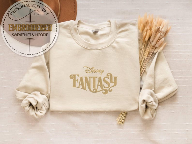 Peut inclure: Sweat-shirt cr&egrave;me avec l'inscription "Disney Fantasy" en lettres dor&eacute;es. Le sweat-shirt a des manches longues et un col rond. L'image comprend &eacute;galement un bouquet de bl&eacute; s&eacute;ch&eacute; et un chapeau.