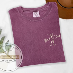 K&ouml;nnte beinhalten: Pflaumenfarbenes Comfort Colors T-Shirt mit dem gestickten Text "Best Dad by Par" und einer Golfspieler-Grafik. Das Shirt ist gefaltet, mit einem braunen Hut im Hintergrund. Das Shirt ist ein personalisiertes Geschenk.