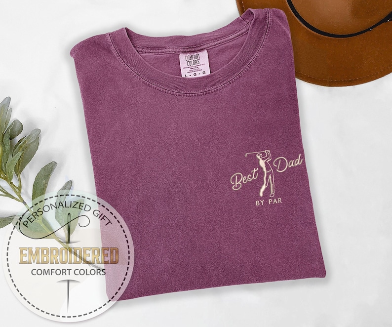 K&ouml;nnte beinhalten: Pflaumenfarbenes Comfort Colors T-Shirt mit dem gestickten Text "Best Dad by Par" und einer Golfspieler-Grafik. Das Shirt ist gefaltet, mit einem braunen Hut im Hintergrund. Das Shirt ist ein personalisiertes Geschenk.