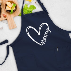 May include: A navy blue apron with a white heart and the name "Nancy" written inside the heart. The apron has a pocket on the front.