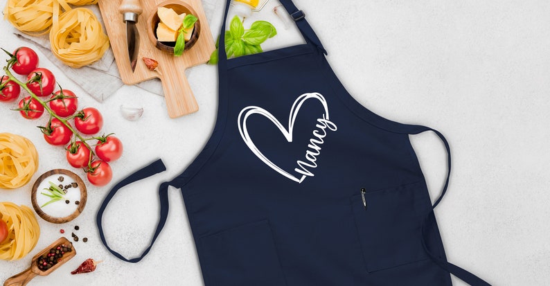 May include: A navy blue apron with a white heart and the name "Nancy" written inside the heart. The apron has a pocket on the front.