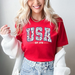 May include: A red t-shirt with the word "USA" in a floral design. The text "EST. 1776" is below the word "USA". The shirt is short-sleeved.