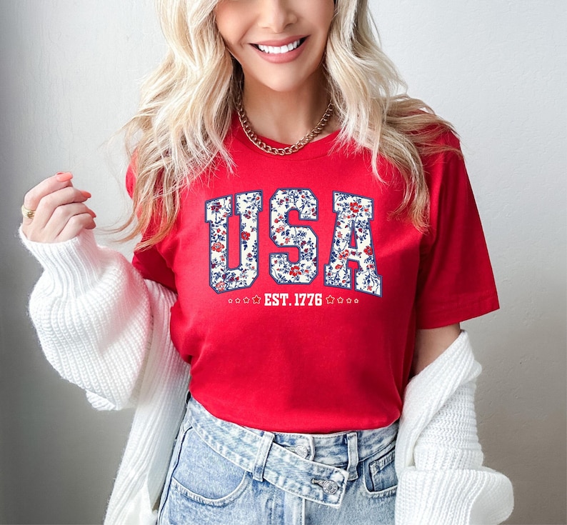 May include: A red t-shirt with the word "USA" in a floral design. The text "EST. 1776" is below the word "USA". The shirt is short-sleeved.