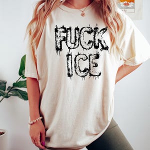 May include: Cream-colored t-shirt with the text "FUCK ICE" in dripping black letters. The shirt is a relaxed fit and has short sleeves. The model is wearing dark green biker shorts.