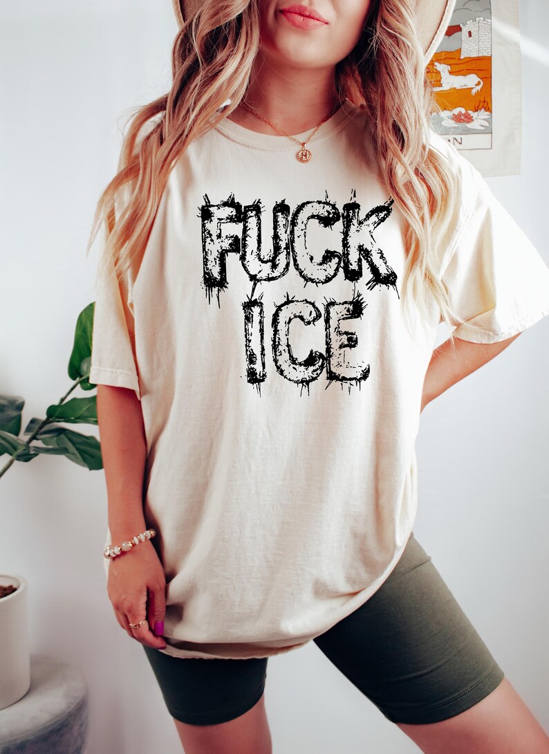 May include: Cream-colored t-shirt with the text "FUCK ICE" in dripping black letters. The shirt is a relaxed fit and has short sleeves. The model is wearing dark green biker shorts.