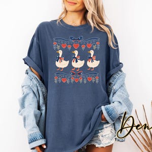 May include: A navy blue t-shirt with a graphic of three white ducks wearing patriotic bows. The design includes red hearts, flowers, and bows. The word "Denim" is written in a cursive font on the bottom right.