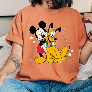 May include: A light orange t-shirt with a cartoon image of Mickey Mouse and Pluto. Mickey Mouse is wearing red shorts and white gloves. Pluto is a yellow dog with a red collar.