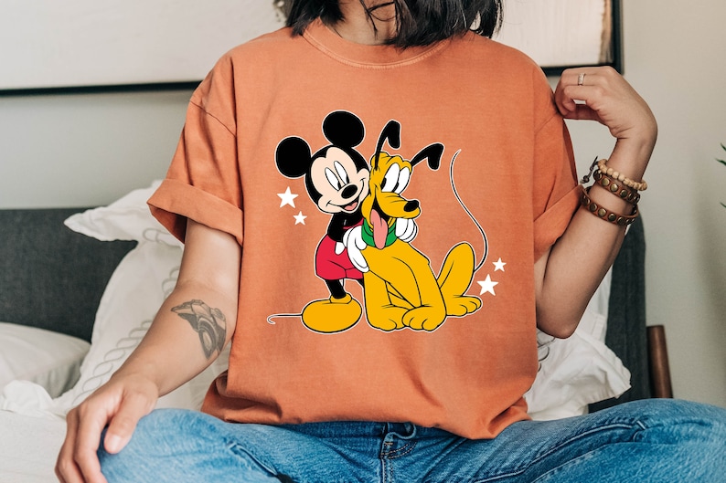 May include: A light orange t-shirt with a cartoon image of Mickey Mouse and Pluto. Mickey Mouse is wearing red shorts and white gloves. Pluto is a yellow dog with a red collar.