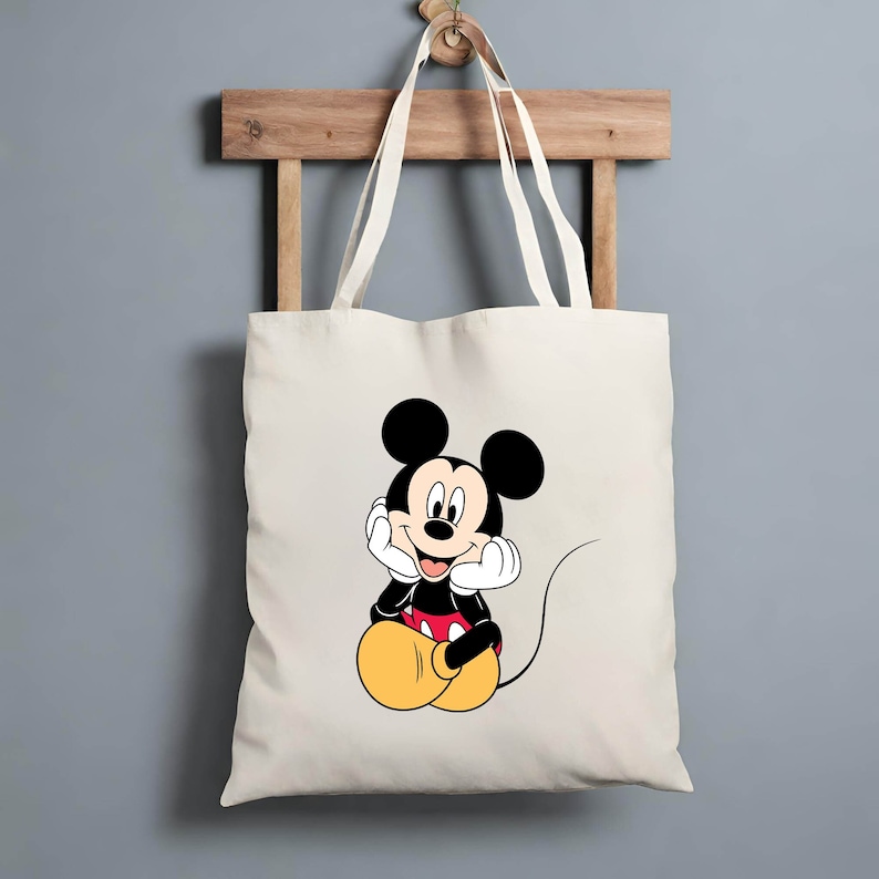 K&ouml;nnte beinhalten: Eine wei&szlig;e Canvas-Tragetasche mit einem Cartoon-Bild von Mickey Mouse, der mit den H&auml;nden am Kinn sitzt. Mickey Mouse tr&auml;gt seine charakteristischen roten Shorts und gelben Schuhe.
