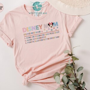 May include: Pink t-shirt with a graphic design that says "Disney Mom" with a cartoon image of Minnie Mouse. The text below the graphic says "(N)/DIZ-NEE MAM" and lists three reasons why the person is a Disney Mom: "1. Runs on Starbucks and Pixie Dust", "2. Magic Coordinator", and "3. Practically Perfect in Every Way".