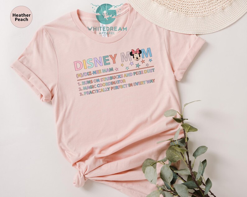 May include: Pink t-shirt with a graphic design that says "Disney Mom" with a cartoon image of Minnie Mouse. The text below the graphic says "(N)/DIZ-NEE MAM" and lists three reasons why the person is a Disney Mom: "1. Runs on Starbucks and Pixie Dust", "2. Magic Coordinator", and "3. Practically Perfect in Every Way".