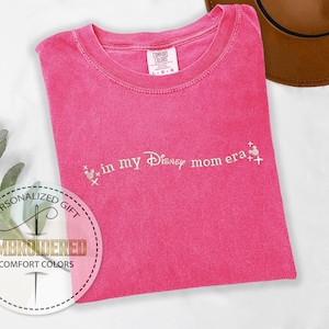 May include: Pink t-shirt with the text "in my Disney mom era" embroidered in white with a small Mickey Mouse graphic.