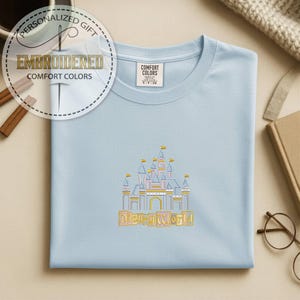 May include: Light blue t-shirt with an embroidered Disney World castle design. The shirt features the text "Disney World" in a gold color. The shirt also has the text "Embroidered Comfort Colors" in a circular design.