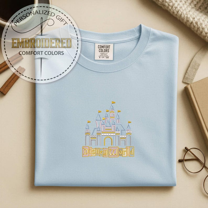 May include: Light blue t-shirt with an embroidered Disney World castle design. The shirt features the text "Disney World" in a gold color. The shirt also has the text "Embroidered Comfort Colors" in a circular design.