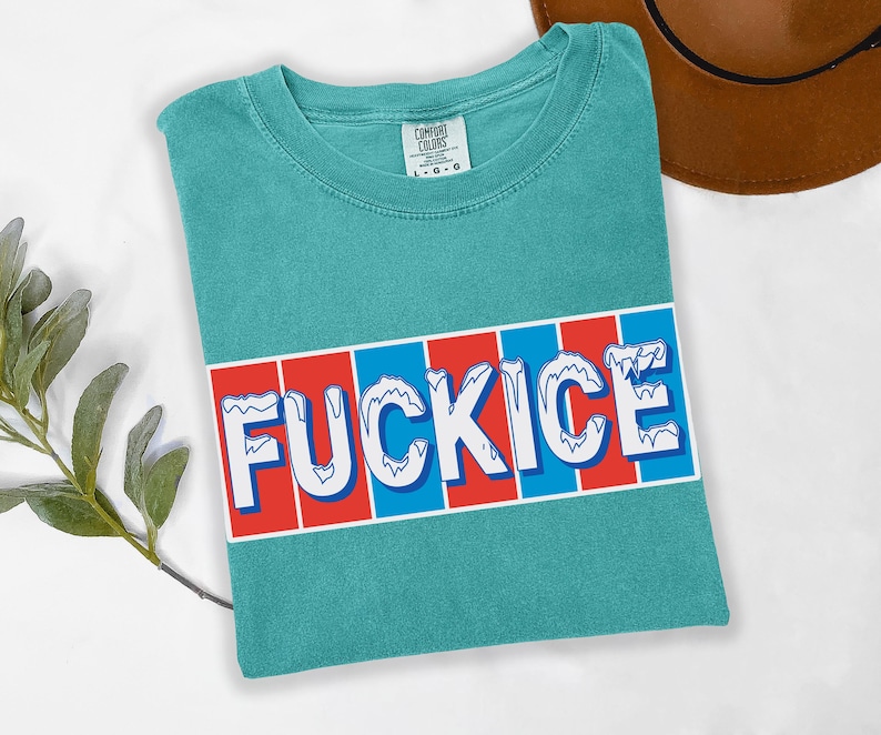 以下が含まれることがあります： ターコイズ色のTシャツ。白で「FUCKICE」の文字が青い縁取りで描かれ、赤と青のストライプの背景に配置されています。Tシャツは折りたたまれており、Comfort Colorsのタグが付いています。