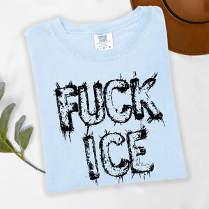 May include: Light blue t-shirt with the words "FUCK ICE" in black, distressed lettering. The shirt is a Comfort Colors brand and is folded on a white surface. A brown hat is visible in the background.