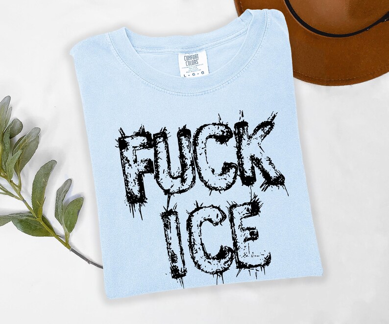 May include: Light blue t-shirt with the words "FUCK ICE" in black, distressed lettering. The shirt is a Comfort Colors brand and is folded on a white surface. A brown hat is visible in the background.