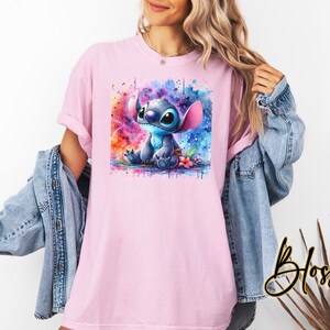 Pu&ograve; includere: Una maglietta rosa chiaro con un'illustrazione ad acquerello del personaggio Stitch di Lilo & Stitch. L'opera d'arte mostra Stitch in blu con orecchie rosa, su uno sfondo vibrante e colorato. La maglietta ha maniche corte.