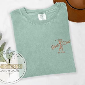 K&ouml;nnte beinhalten: Salbeigr&uuml;nes Comfort Colors T-Shirt mit gesticktem Design. Das Design zeigt einen Golfspieler und die Worte "Best Dad by Par" in kupferfarbenem Garn. Das Shirt ist gefaltet und auf einer wei&szlig;en Oberfl&auml;che ausgestellt.