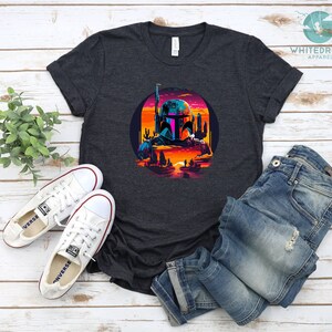Pu&ograve; includere: Una t-shirt grigio scuro melange con una grafica retr&ograve; di Boba Fett in un paesaggio desertico. La grafica presenta un tramonto, cactus e una citt&agrave; futuristica.
