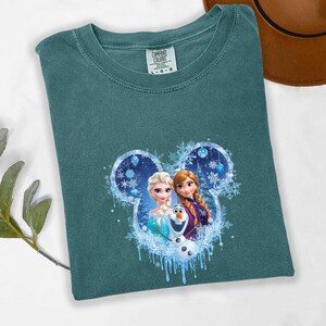 Puede incluir: Camiseta de color verde azulado con un gr&aacute;fico de Elsa, Anna y Olaf de la pel&iacute;cula Frozen, dentro de un contorno de oreja de Mickey Mouse con detalles de copos de nieve. La camiseta es de material suave.