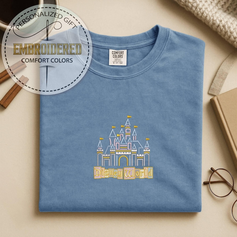 May include: A light blue t-shirt with an embroidered design of a castle and the words "Disney World" in gold. The shirt also has the words "Embroidered" and "Comfort Colors" in a circular design.
