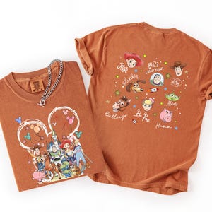May include: Two rust-colored t-shirts with Toy Story graphics. One shirt features a Mickey Mouse-shaped design with characters and balloons. The other shirt displays character faces and names like Buzz Lightyear and Woody. A silver chain necklace rests on the folded shirt.
