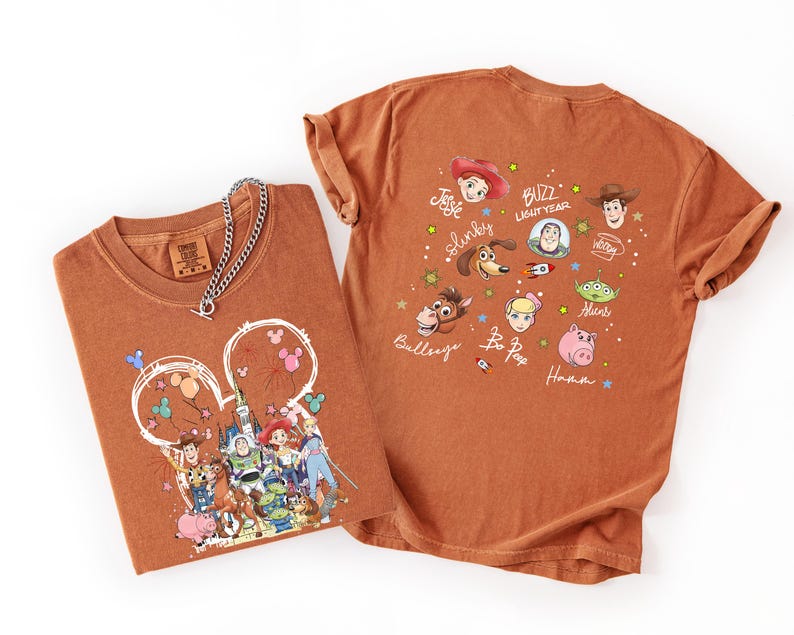 May include: Two rust-colored t-shirts with Toy Story graphics. One shirt features a Mickey Mouse-shaped design with characters and balloons. The other shirt displays character faces and names like Buzz Lightyear and Woody. A silver chain necklace rests on the folded shirt.