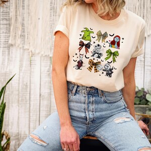 May include: A cream-colored t-shirt featuring a colorful graphic of characters and bows from a popular animated film. The design includes images of a green bow, a skeleton, and a character with red hair. The shirt is made of a soft material.