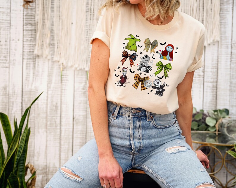 May include: A cream-colored t-shirt featuring a colorful graphic of characters and bows from a popular animated film. The design includes images of a green bow, a skeleton, and a character with red hair. The shirt is made of a soft material.