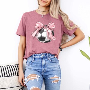 May include: A mauve t-shirt with a graphic of a soccer ball with a pink bow on it.