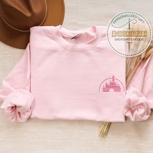 May include: Pink sweatshirt with a pink embroidered image of the Disney castle on the front. The sweatshirt is laid flat on a light brown surface.