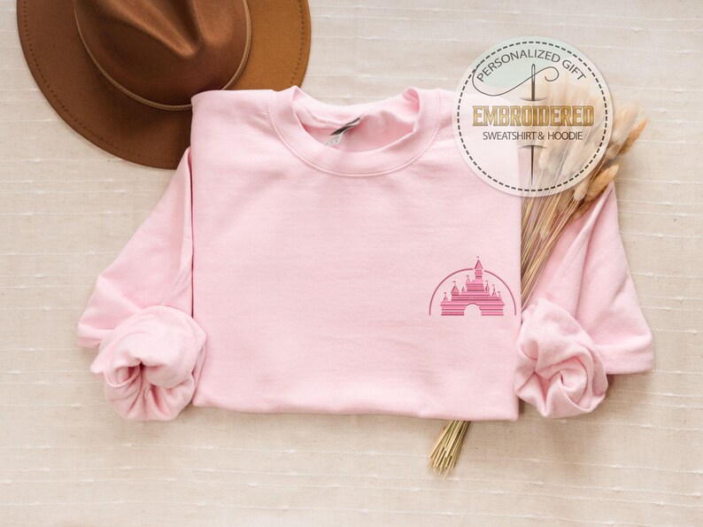 May include: Pink sweatshirt with a pink embroidered image of the Disney castle on the front. The sweatshirt is laid flat on a light brown surface.