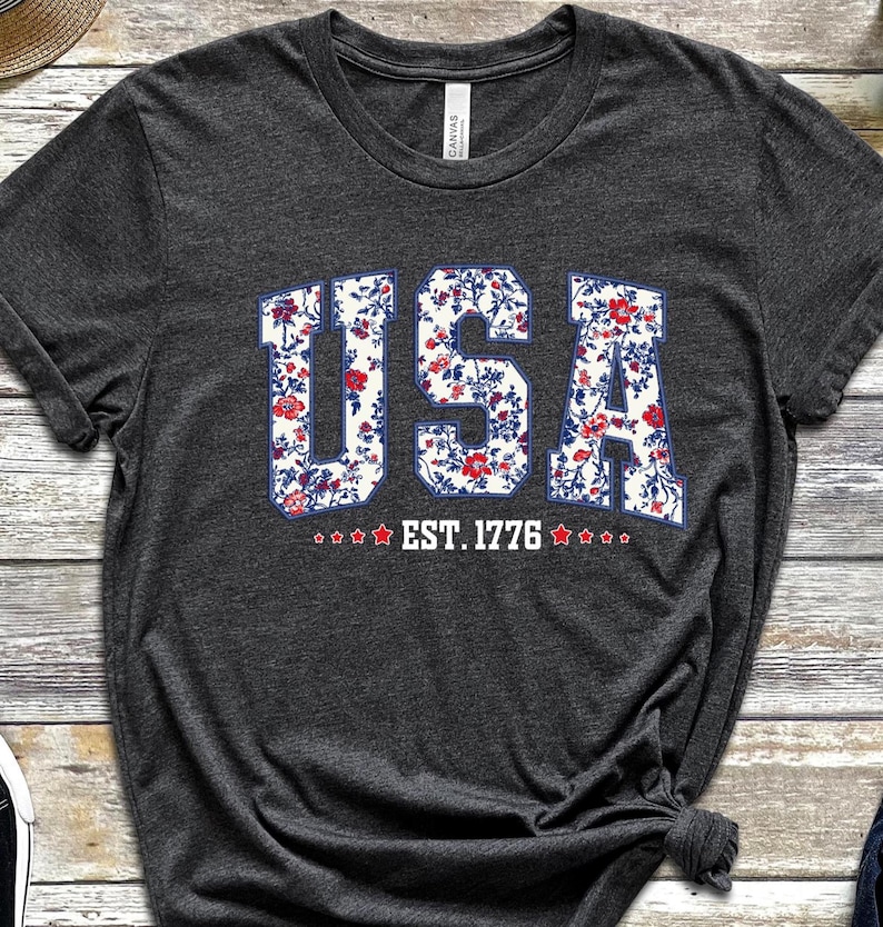 May include: Dark gray t-shirt with the word "USA" in a floral pattern, outlined in blue. Below the word "USA" is the text "EST. 1776" with star accents. The shirt is made of a soft material.