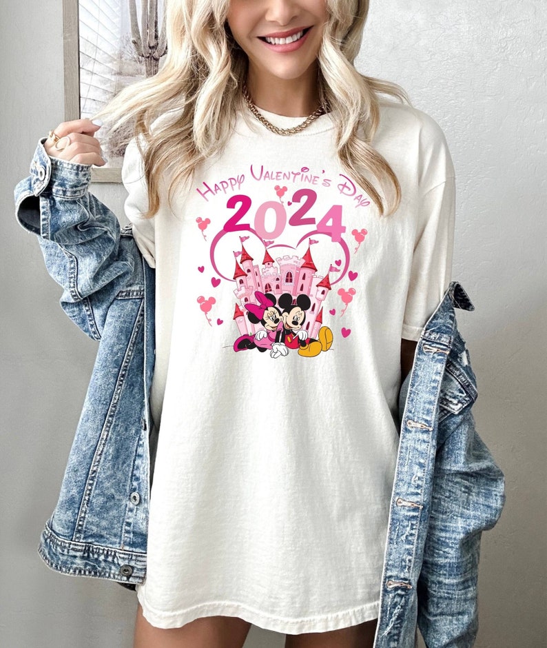 May include: White t-shirt with a pink graphic of Mickey Mouse and Minnie Mouse in front of a pink castle. The graphic also includes the text "Happy Valentine's Day 2024" with pink hearts.