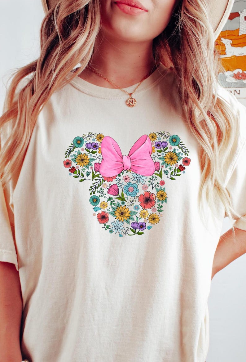 Op de afbeelding: Cr&egrave;mekleurig T-shirt met een bloemen Minnie Mouse-ontwerp. Het ontwerp bevat een roze strik en een verscheidenheid aan kleurrijke bloemen in geel, blauw, rood en paars, wat een vrolijke en grillige esthetiek cre&euml;ert.