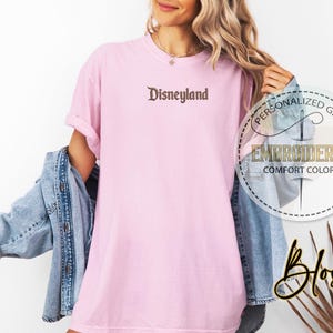 以下が含まれることがあります： 薄いピンク色のTシャツ。茶色の筆記体フォントで「Disneyland」の文字が刺繍されています。このシャツはパーソナライズされたギフトコレクションの一部で、快適な色合いの素材で作られています。デニムジャケットがシャツの上に着用されています。