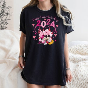 May include: Black t-shirt with a pink graphic of Mickey Mouse and Minnie Mouse in front of a pink castle. The text "Happy Valentine's Day 2024" is above the characters.