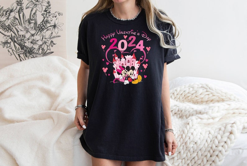 May include: Black t-shirt with a pink graphic of Mickey Mouse and Minnie Mouse in front of a pink castle. The text "Happy Valentine's Day 2024" is above the characters.