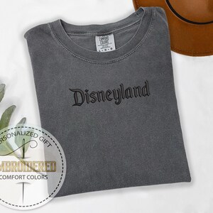 以下が含まれることがあります： 黒い筆記体で「Disneyland」と刺繍されたグレーのコンフォートカラーズTシャツ。 シャツは折りたたまれ、白い表面に置かれています。 背景には茶色の帽子が見えます。 シャツには「Comfort Colors」と書かれたタグが付いています。