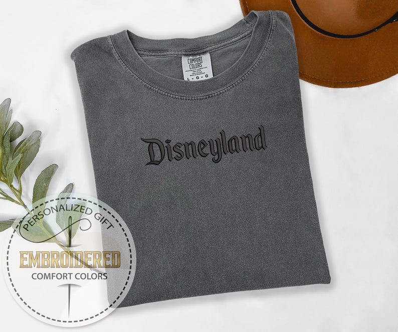 以下が含まれることがあります： 黒い筆記体で「Disneyland」と刺繍されたグレーのコンフォートカラーズTシャツ。 シャツは折りたたまれ、白い表面に置かれています。 背景には茶色の帽子が見えます。 シャツには「Comfort Colors」と書かれたタグが付いています。