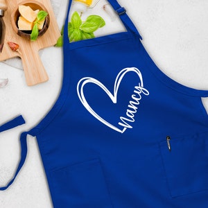 May include: A blue apron with a white heart and the name "Nancy" written inside. The apron has a pocket on the front.