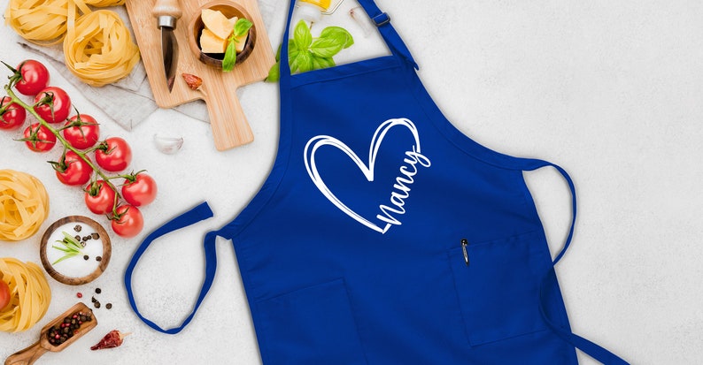 May include: A blue apron with a white heart and the name "Nancy" written inside. The apron has a pocket on the front.