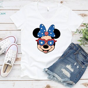 May include: White V-neck t-shirt with a red, white, and blue Minnie Mouse graphic wearing heart-shaped sunglasses. The graphic features a red bow with white stars and a red, white, and blue bow. The shirt is laying on a white wooden surface with a pair of white Converse shoes and a pair of blue denim shorts.
